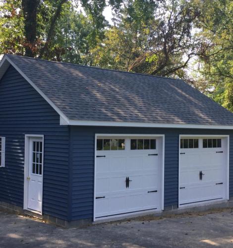 2 car garage with blue siding, 2 white garage doors, a side entry door, windows with white shutters, white trim, and a gray shingle roof.