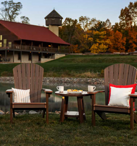 2 dark brown poly Adirondack chairs with white and red pillows sit next to a matching poly side table with food, there's a creek and a large cabin behind them.
