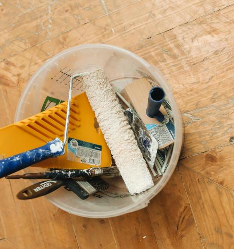 Bucket sits on the floor with a paint roller, paint brushes, and other painting supplies inside.