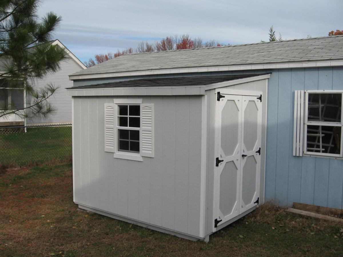 Light gray Lean-To Shed with white trim, a window with white shutters, and double doors with white trim is placed alongside a blue building.
