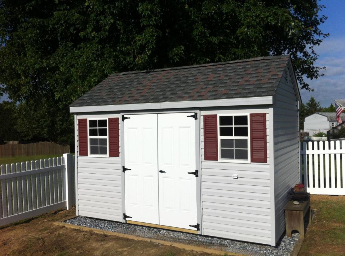 Cape Cottage Shed with light gray vinyl siding, white double doors, 2 windows with red shutters, and dark shingle roofing sits in a fenced-in backyard.