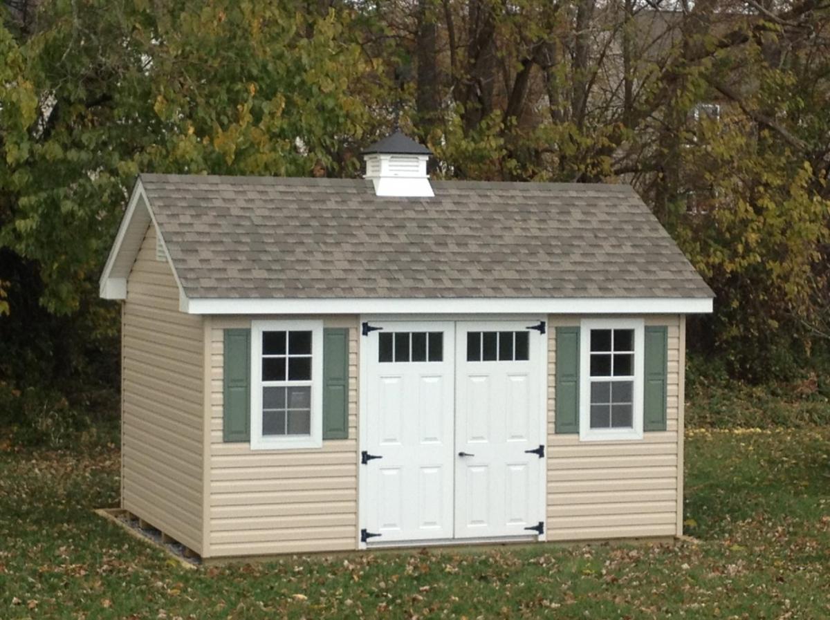 Classic Garden Cottage Shed with tan vinyl siding, white double doors, 2 white windows with green shutters, brown shingle roofing, and a white cupola.