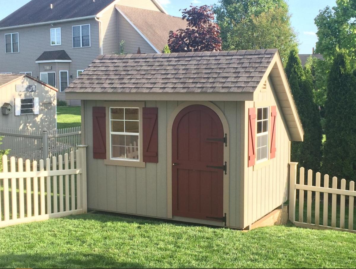 Classic Garden Quaker Shed with tan siding, a red door, 2 windows with red shutters, and a brown asphalt roof sits alongside fencing in a backyard.