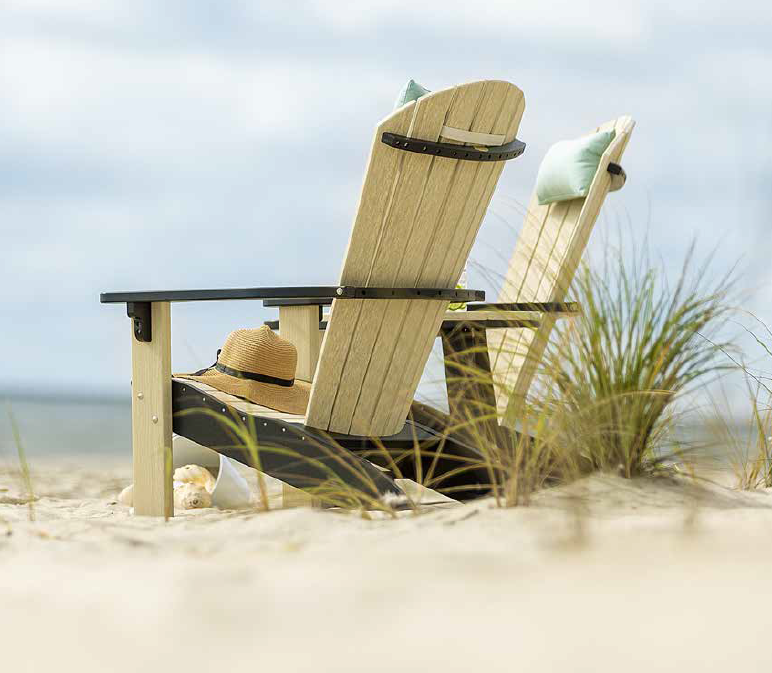 2 light colored poly Adirondack chairs with black accents and pillow head rests sit in the sand on a beach overlooking the water.