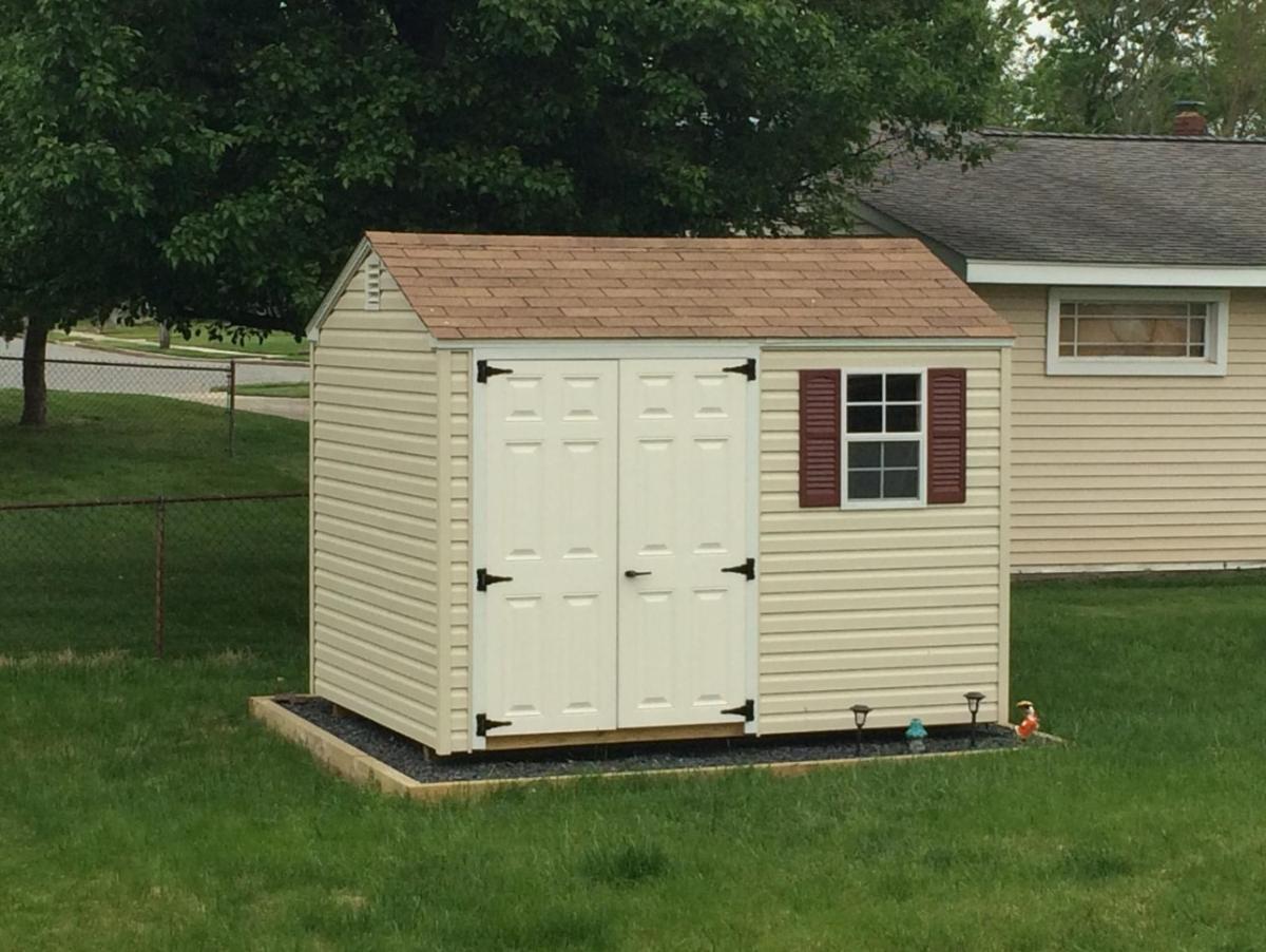Salem Shed with light yellow vinyl siding, brow roofing, white double doors, and a window with red shutters sits in a backyard.